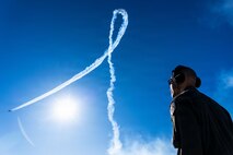 Gen. Spain looks up to the sky as a Thunderbird is flying in the far distance, leaving a white trail that passes the sun.