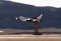 A Thunderbird aircraft flies low on the flightline with the air control tower and mountains blurred in the background.