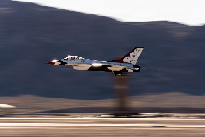 A Thunderbird aircraft flies low on the flightline with the air control tower and mountains blurred in the background.