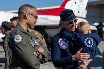 Gen. Adrian Spain and Lt Col Tyler Clark, carrying a child, are smiling looking off camera with a Thunderbird aircraft and bystanders in the background.