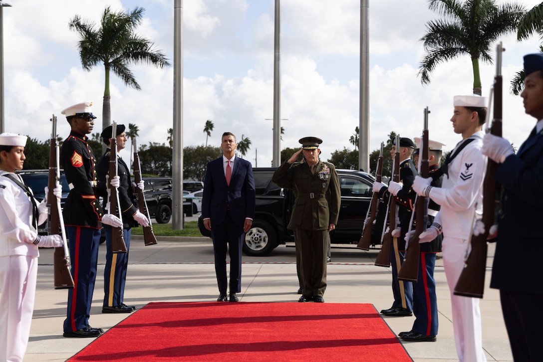 A man in a brown military uniform and a man in a dark blue suit stand at the top of a red carpet that is flanked by a miliary honor guard.