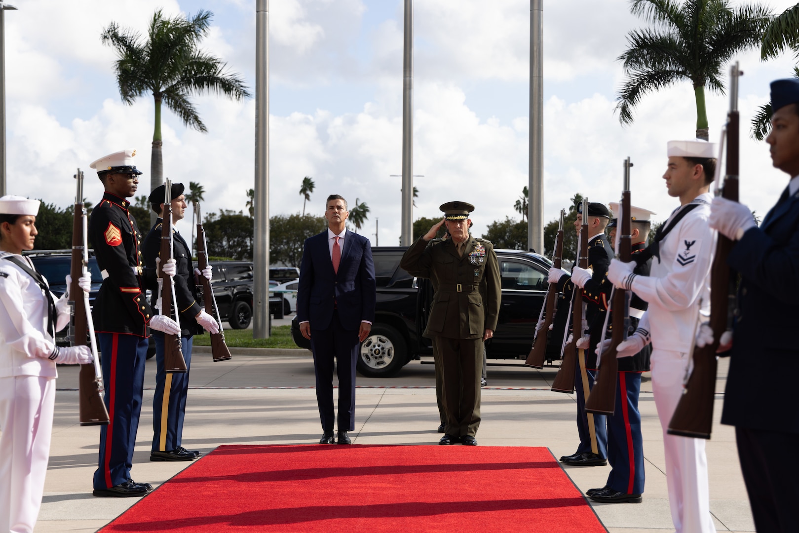 A man in a brown military uniform and a man in a dark blue suit stand at the top of a red carpet that is flanked by a miliary honor guard.
