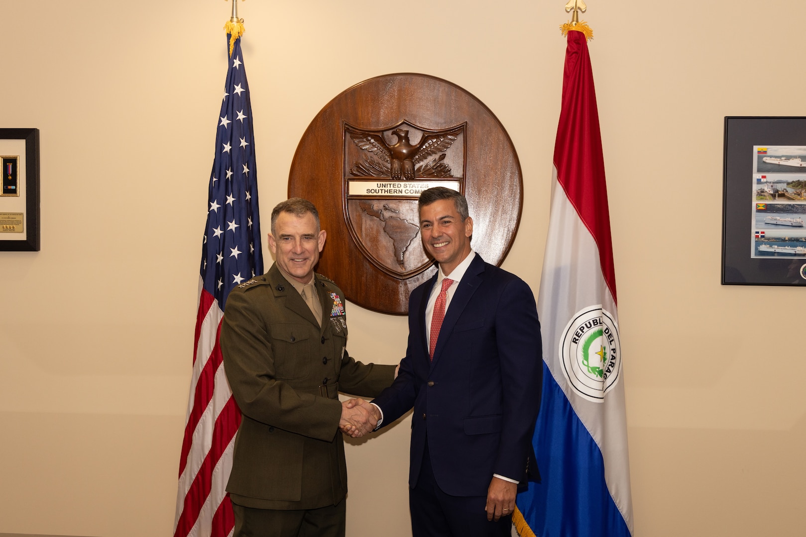 A man in a brown military uniform shakes hands with a man in a dark blue suit in front of 2 flags.