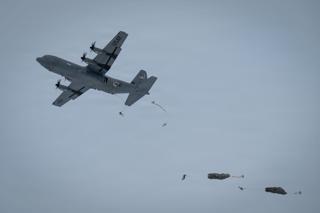 A low angle of an aircraft flying through the sky while four soldiers with parachutes jump out one at a time.