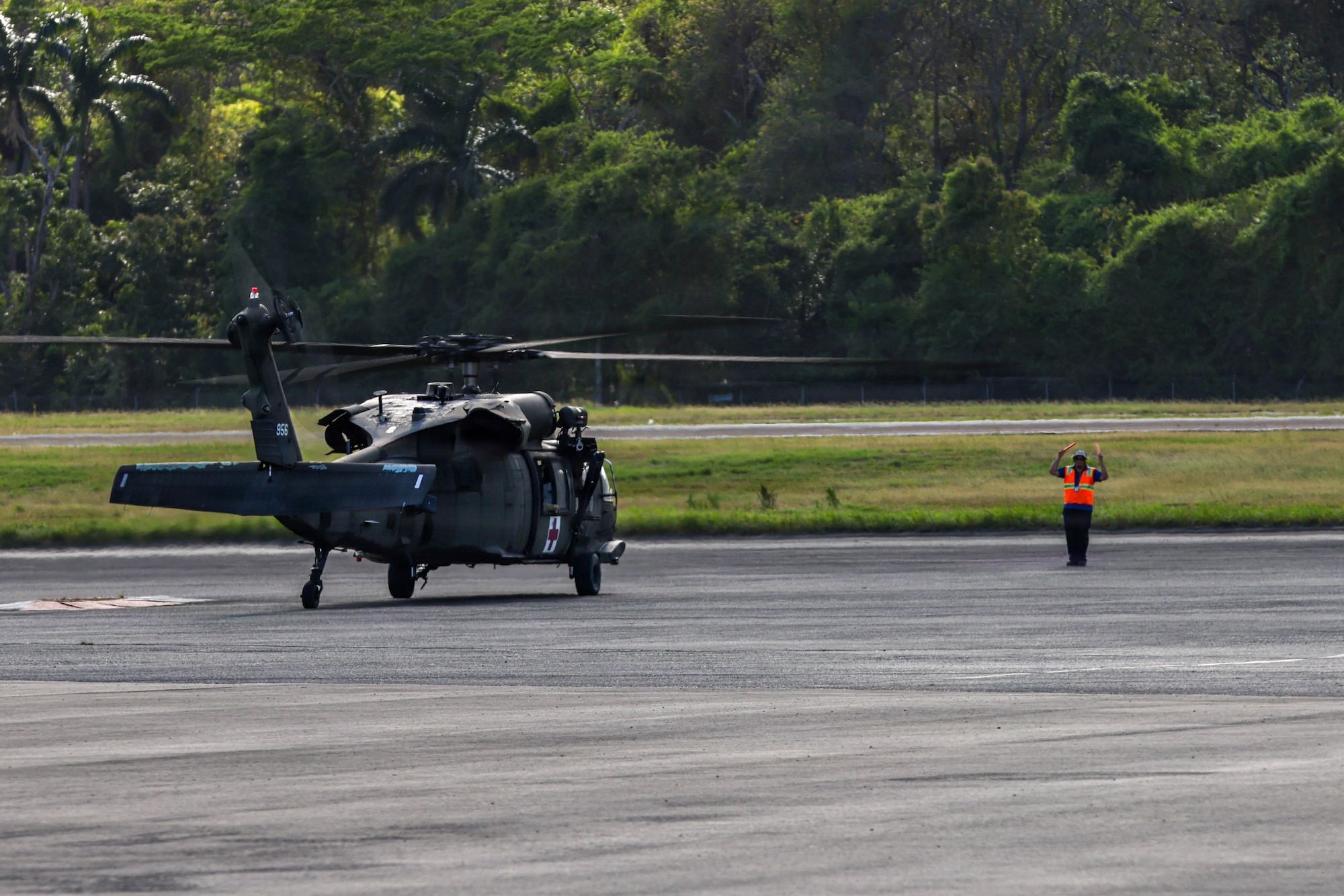An airfield worker wearing an orange vest guides a military helicopter to a parking spot.