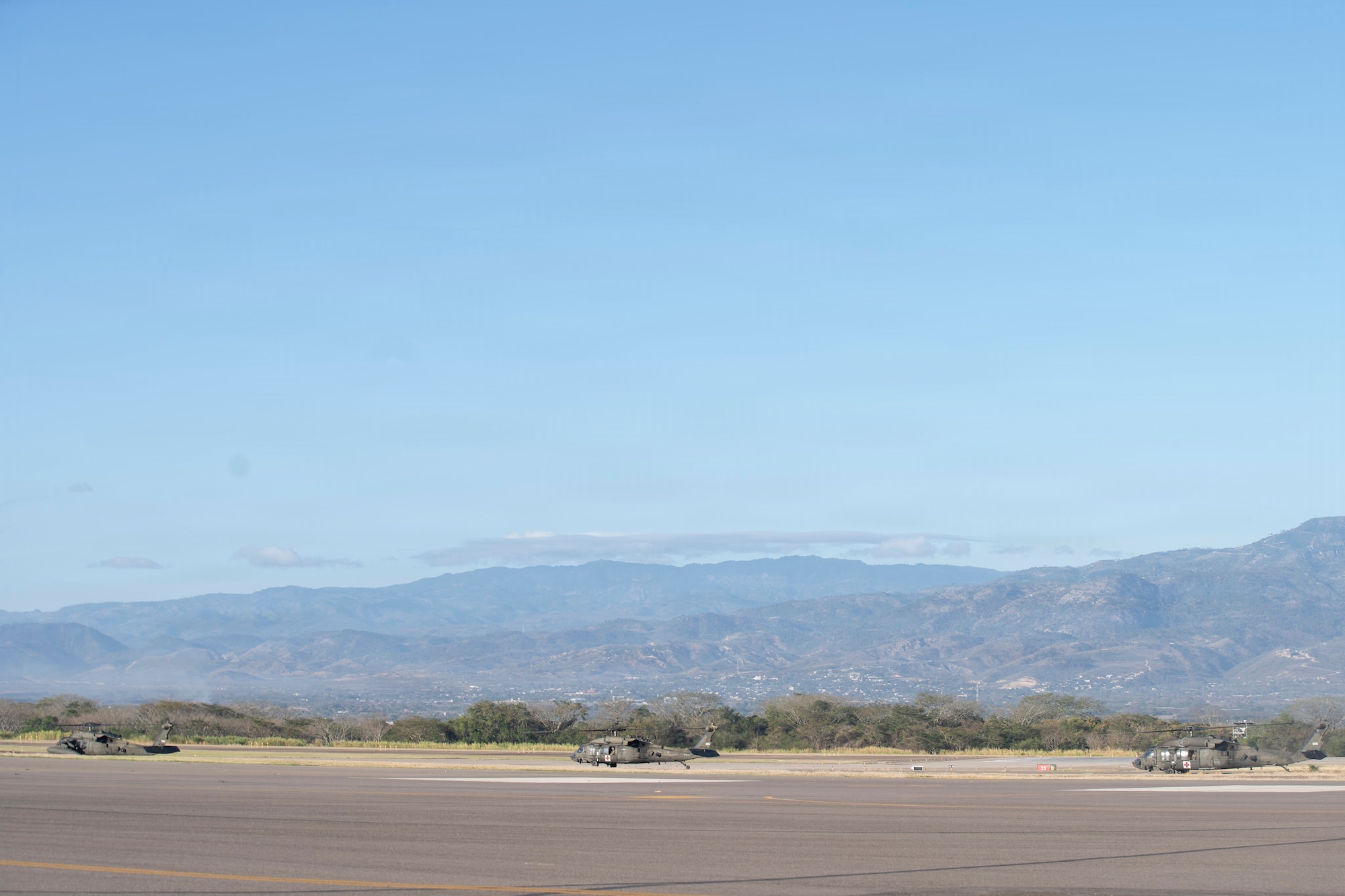 3 military helicopters idle on the flight line with mountains in the background.