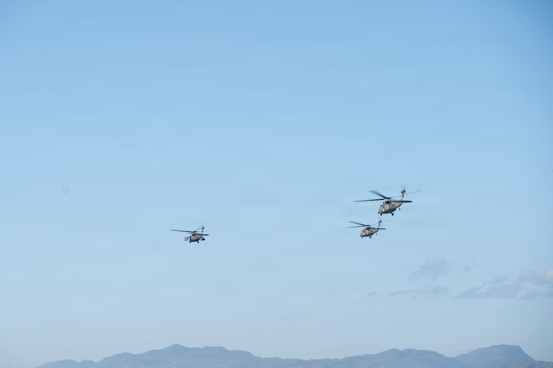 3 military helicopters fly in the sky over some mountains in the background.