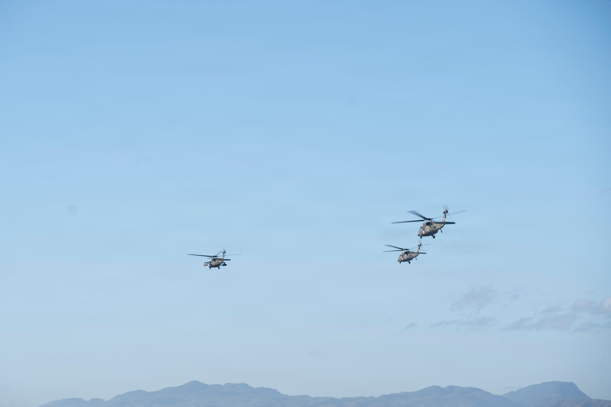 3 military helicopters fly in the sky over some mountains in the background.