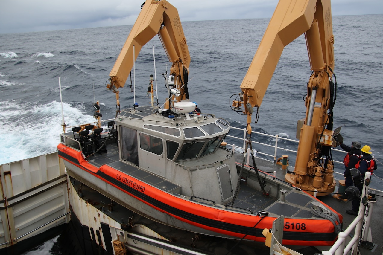 A small boat is lifted from the deck of a larger ship by people in military uniforms and hard hats using two hydraulic machines. The ship is sailing in the ocean.