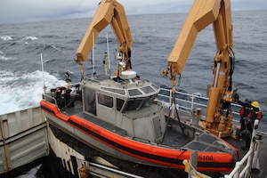 A small boat is lifted from the deck of a larger ship by people in military uniforms and hard hats using two hydraulic machines. The ship is sailing in the ocean.