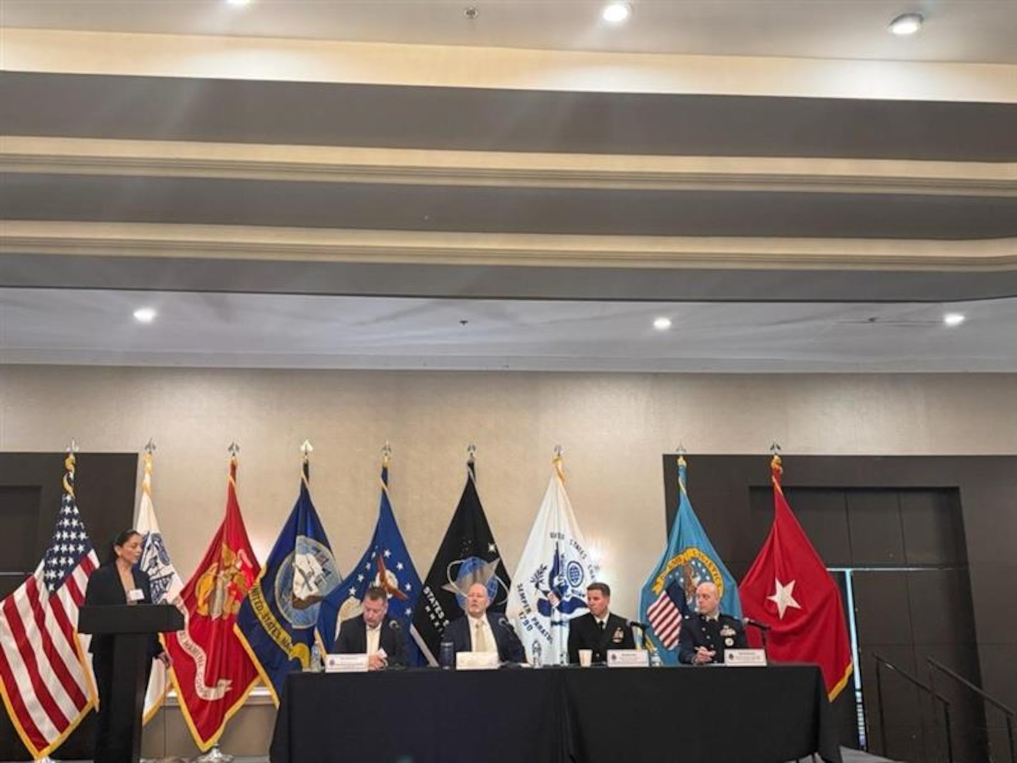 woman stands at podium, four men sit behind table on stage