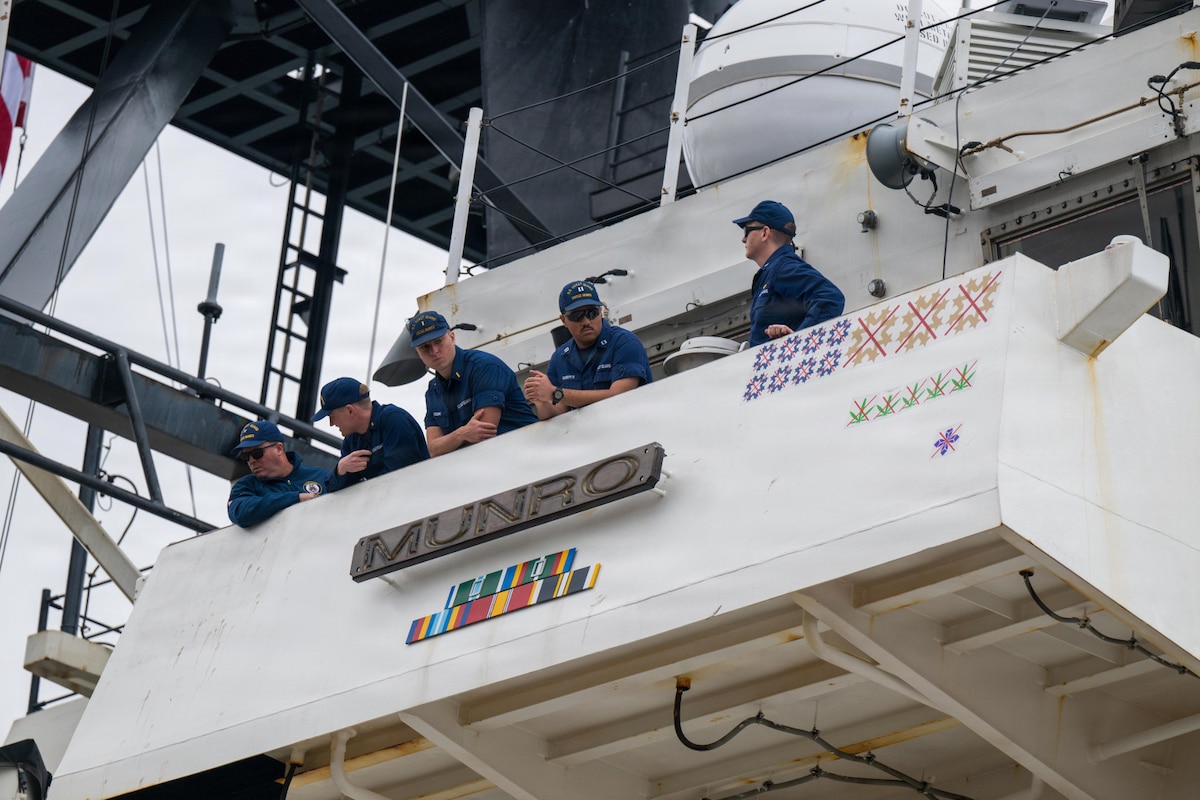 Five men in military uniforms stand looking out from the bridge of a large ship. There is a sign on the ship that reads “Munro.”