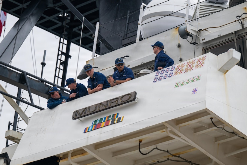 Five men in military uniforms stand looking out from the bridge of a large ship. There is a sign on the ship that reads “Munro.”