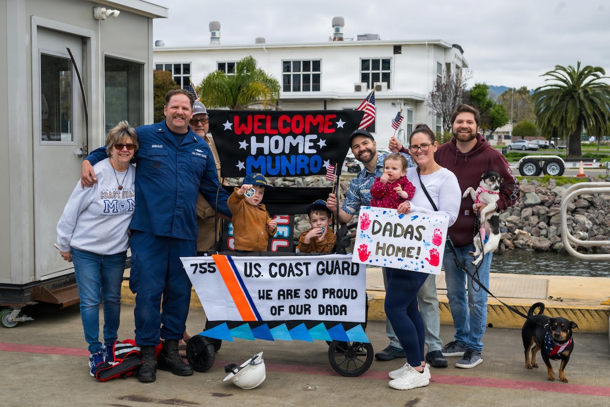 A man in a military uniform poses for a photo with his family on a dock. Several of the people are holding signs and small American flags. Two children in a stroller are holding military challenge coins.