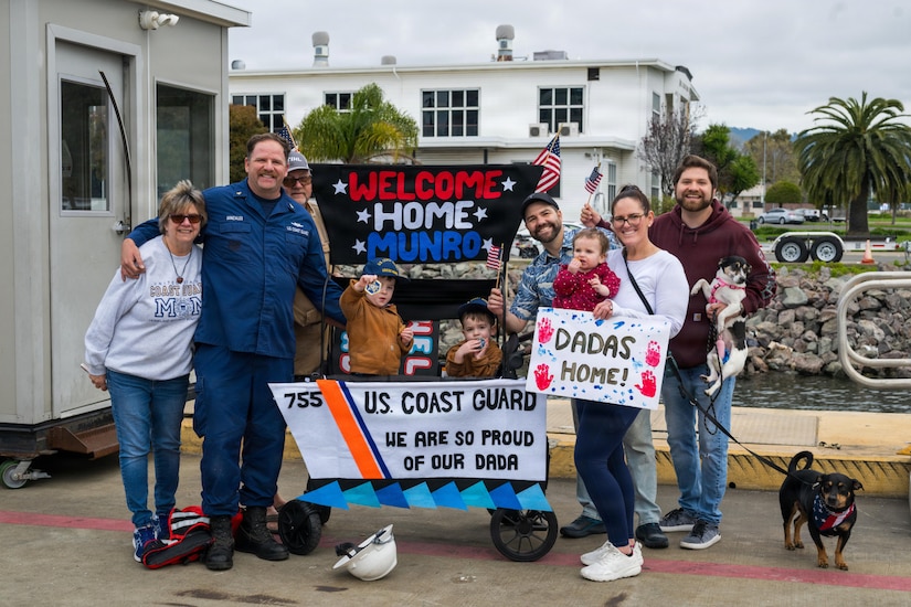 A man in a military uniform poses for a photo with his family on a dock. Several of the people are holding signs and small American flags. Two children in a stroller are holding military challenge coins.