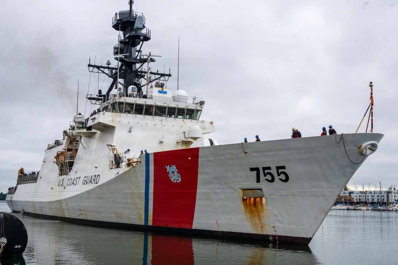 A large white ship pulls into a pier. Several people in military uniforms are manning the lines on the bow of the ship. Painted on the side of the ship is “U.S. Coast Guard.”