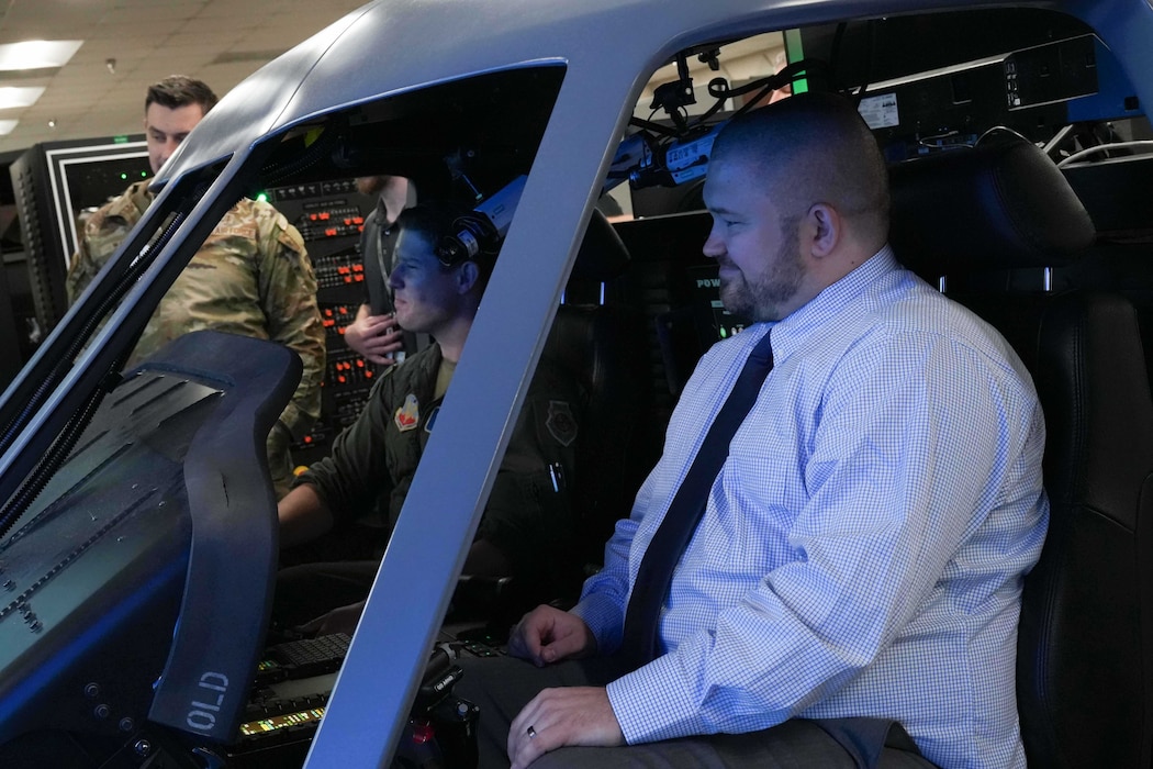 a civilian smiles while sitting inside a helicopter simulator with a uniformed Airman
