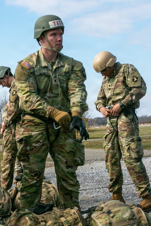A man wearing a camouflage military uniform, rappelling gear and a tactical helmet puts on gloves. The are two other men in similar attire behind him.