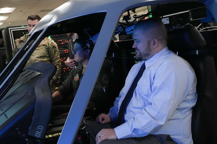 a civilian smiles while sitting inside a helicopter simulator with a uniformed Airman