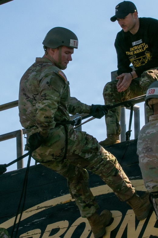 A man wearing a camouflage military uniform, rappelling gear and a tactical helmet prepares to rappel from a wooden tower while holding a rope. There is a man wearing a baseball cap and T-shirt that reads, “Instructor, The Sabalauski Air Assault School,” sitting at the top edge of the rappel tower watching the other man.