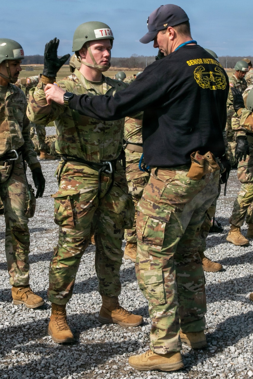 A man wearing a camouflage military uniform, rappelling gear, gloves and a tactical helmet raises his arms as another man inspects his uniform. The other man is wearing a black T-shirt that reads “senior instructor,” a baseball cap, camouflage military uniform pants and boots.