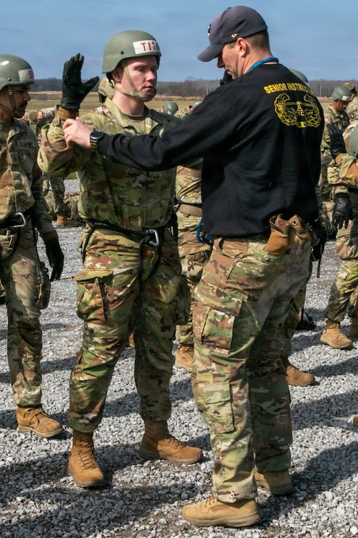 A man wearing a camouflage military uniform, rappelling gear, gloves and a tactical helmet raises his arms as another man inspects his uniform. The other man is wearing a black T-shirt that reads “senior instructor,” a baseball cap, camouflage military uniform pants and boots.