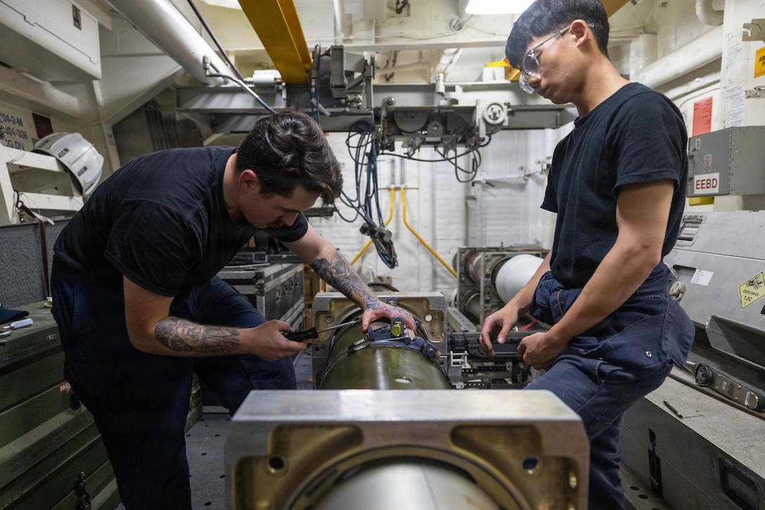 Two sailors in black shirts use tools to band a torpedo in a maintenance room aboard a ship.