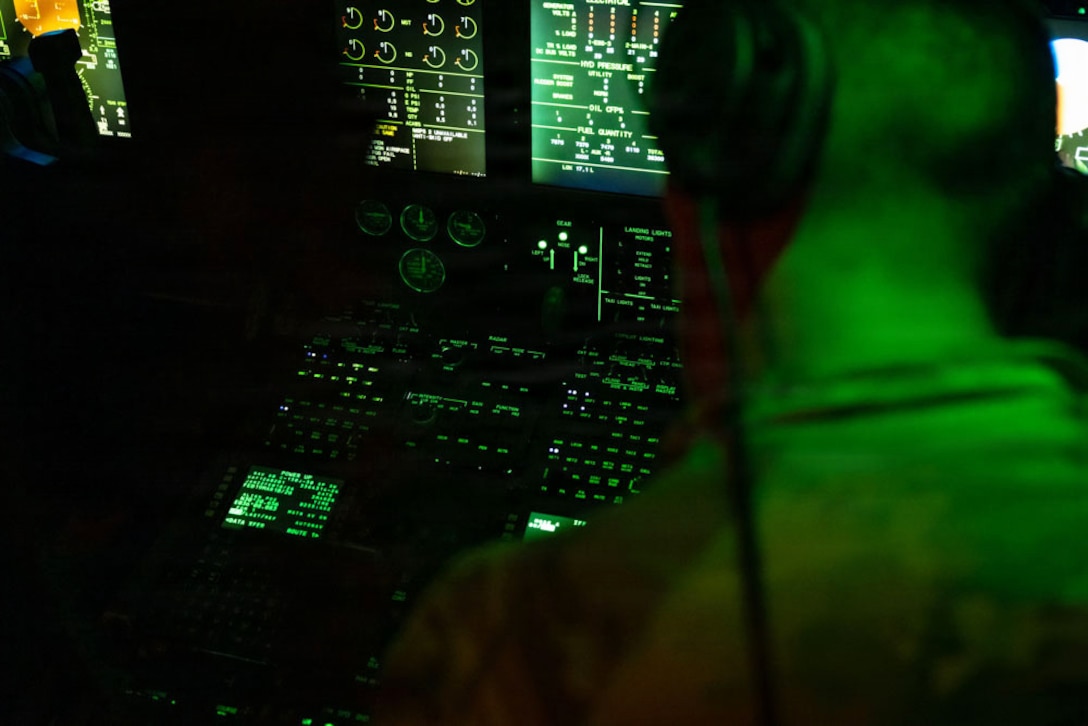 An airman wearing a headset faces a control panel in the dark illuminated by green lights.