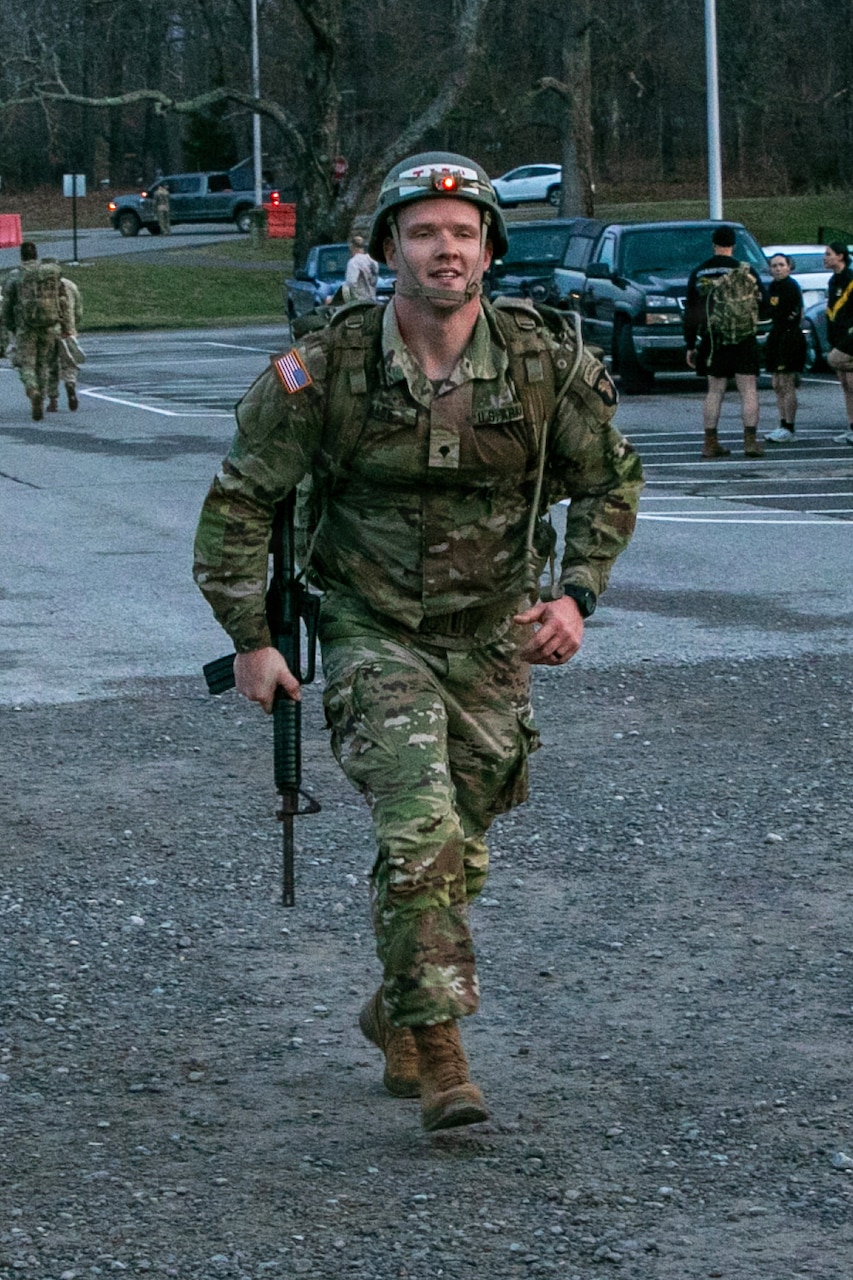 A man wearing a camouflage military uniform, rucksack and a tactical helmet smiles while carrying a rifle as he completes a march.