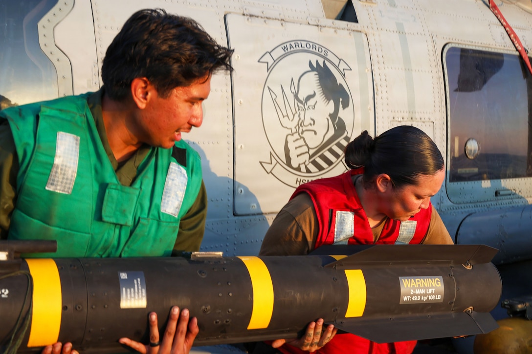 Two sailors in green and red vests hold a missile in front of a helicopter.