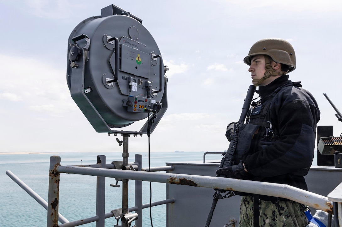 A sailor in a helmet holding a weapon stands watch at the top of a ship at sea against a blue sky.