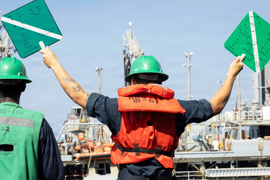 A sailor stands next to a fellow sailor holding green signs toward a ship at sea under a blue sky.