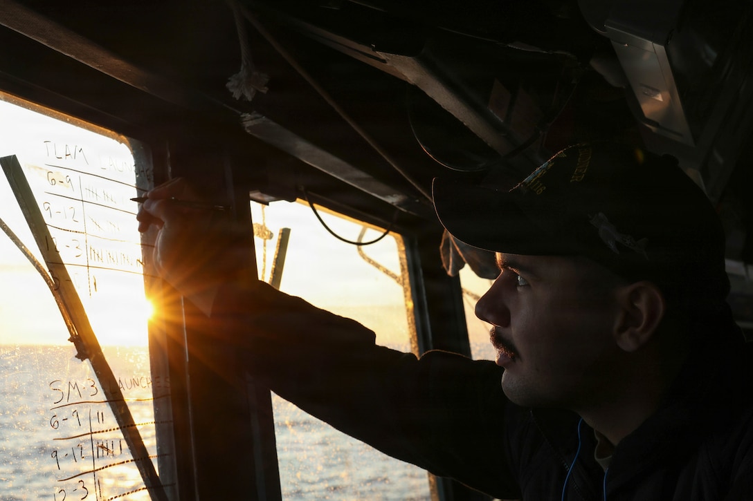 The sun beams through a ship as a sailor writes a tally mark on the window.