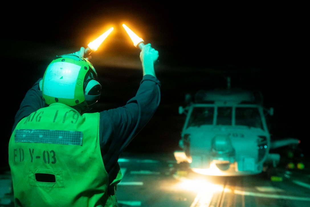 A sailor in a yellow helmet and vest holds two glowing batons in front of a helicopter aboard a ship at sea in the dark.