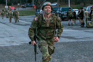 A man wearing a camouflage military uniform, rucksack and a tactical helmet smiles while carrying a rifle as he completes a march.