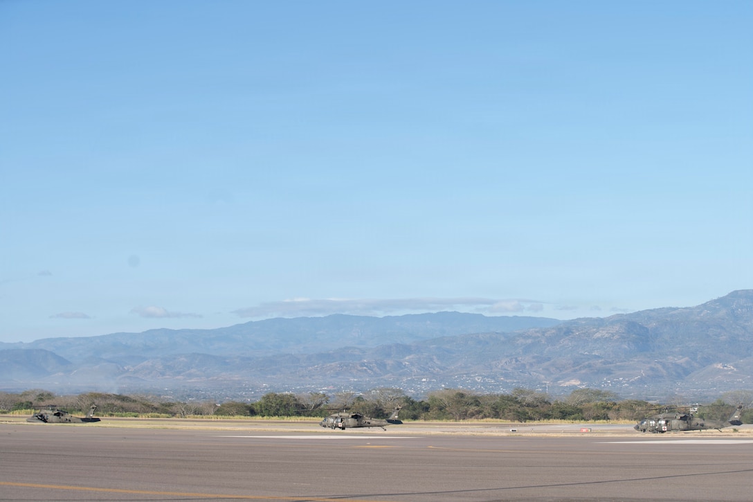3 military helicopters idle on the flight line with mountains in the background.