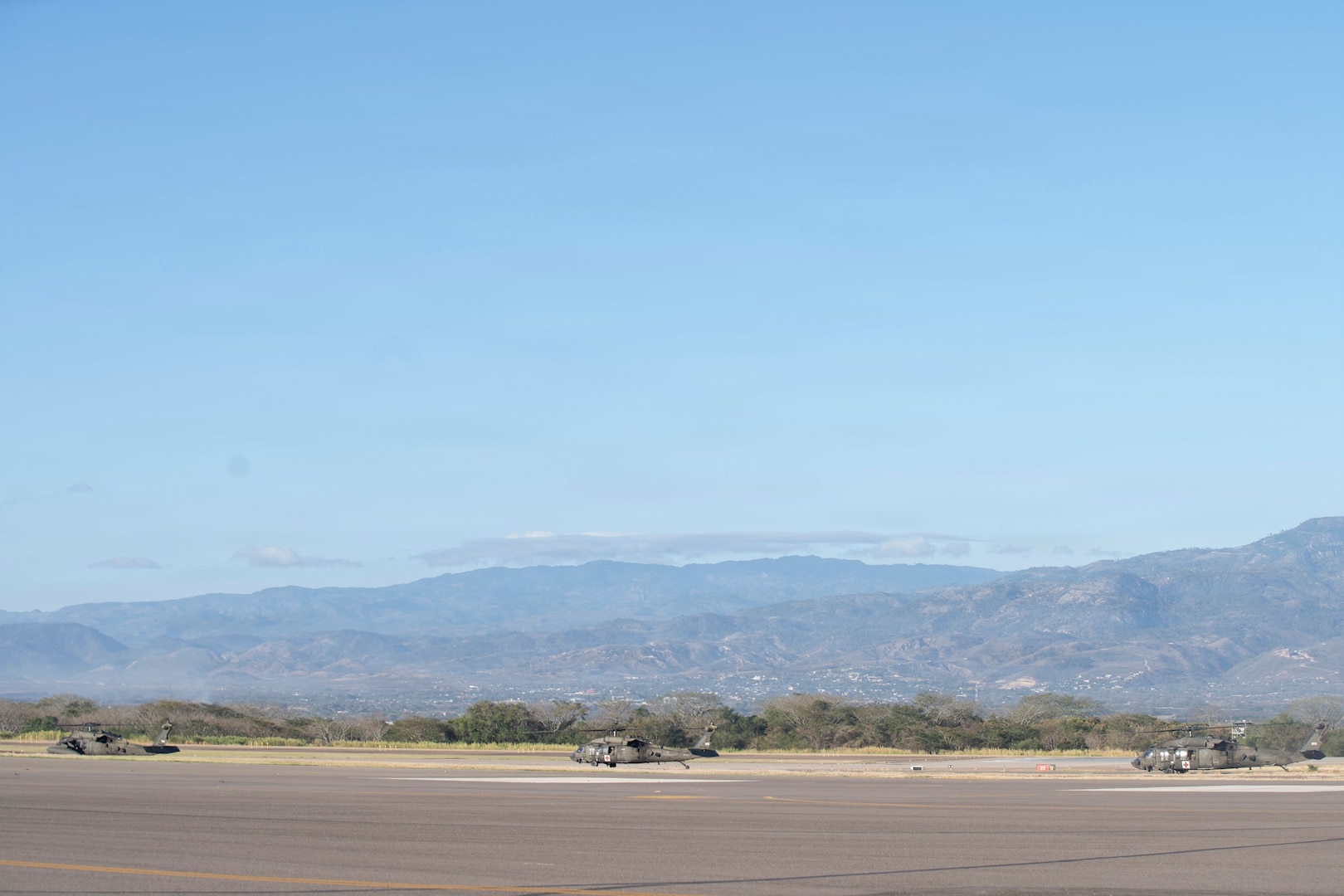 3 military helicopters idle on the flight line with mountains in the background.