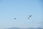 3 military helicopters fly in the sky over some mountains in the background.