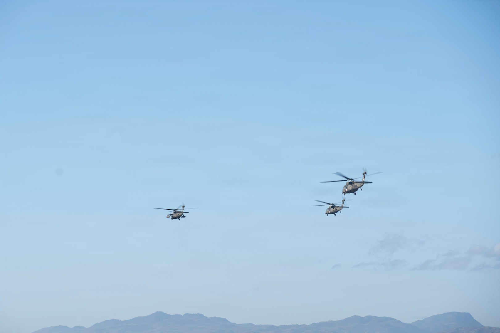 3 military helicopters fly in the sky over some mountains in the background.