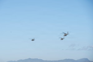 3 military helicopters fly in the sky over some mountains in the background.
