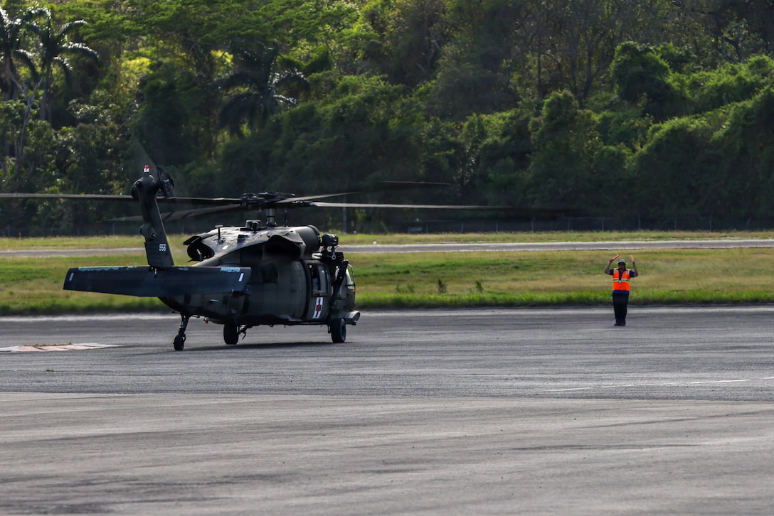An airfield worker wearing an orange vest guides a military helicopter to a parking spot.