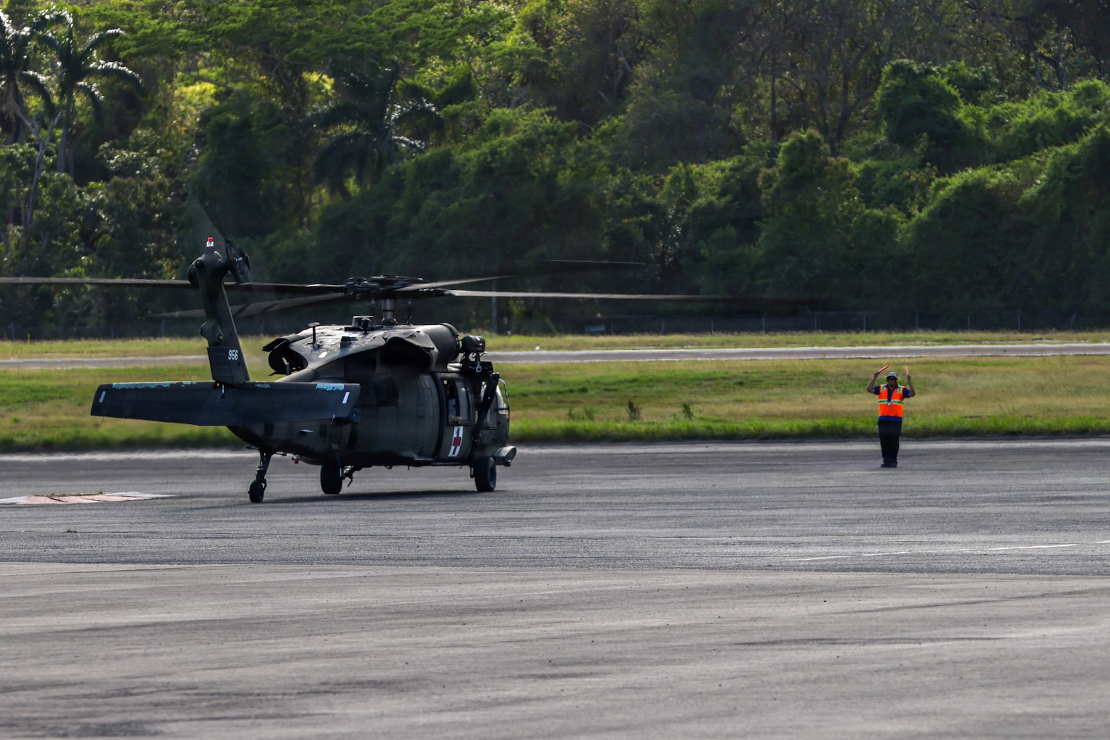 An airfield worker wearing an orange vest guides a military helicopter to a parking spot.