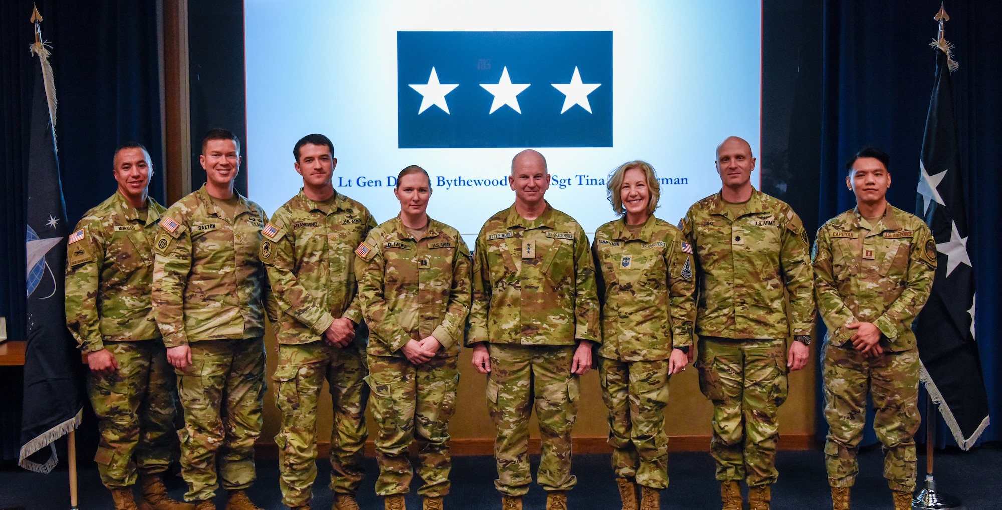 Eight military members stand in front of a projector screen