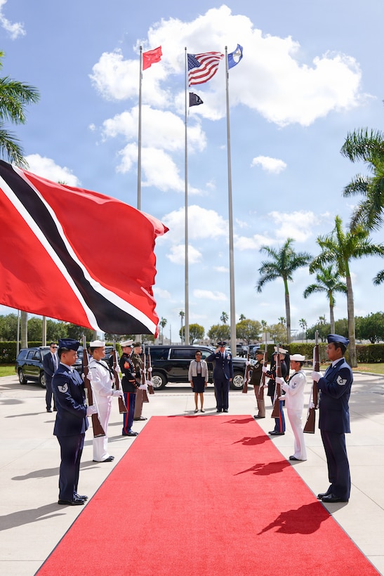 A woman in a white and blue dress and a man in a blue military uniform stand at the end of a red carpet that is flanked by a military honor guard.