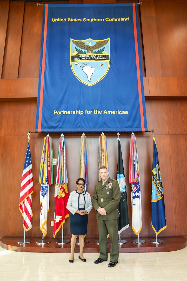 A woman in a white and blue dress shakes hands and poses for a photo with a man in a brown military uniform in front of two flags.