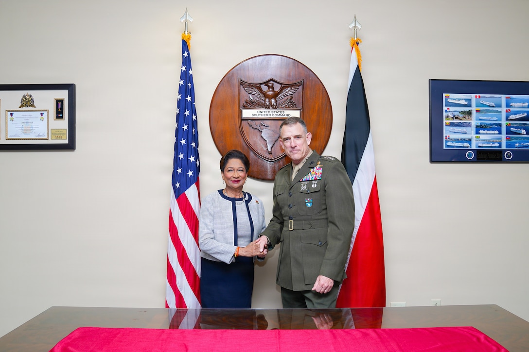 A woman in a white and blue dress shakes hands and poses for a photo with a man in a brown military uniform in front of two flags.