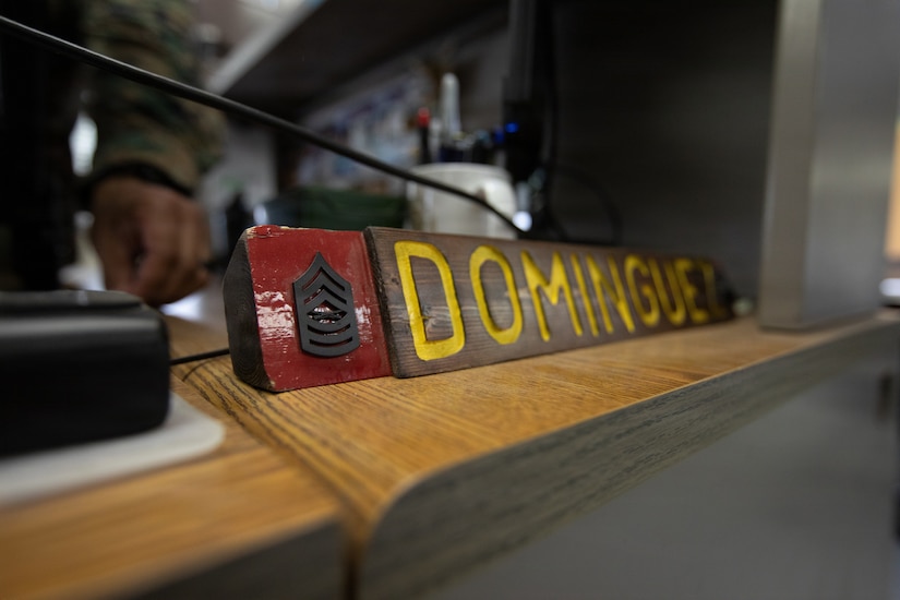 A placard with a military rank and reading "Dominguez" sits on a desk, with a person's hand and lower arm visible in the background.