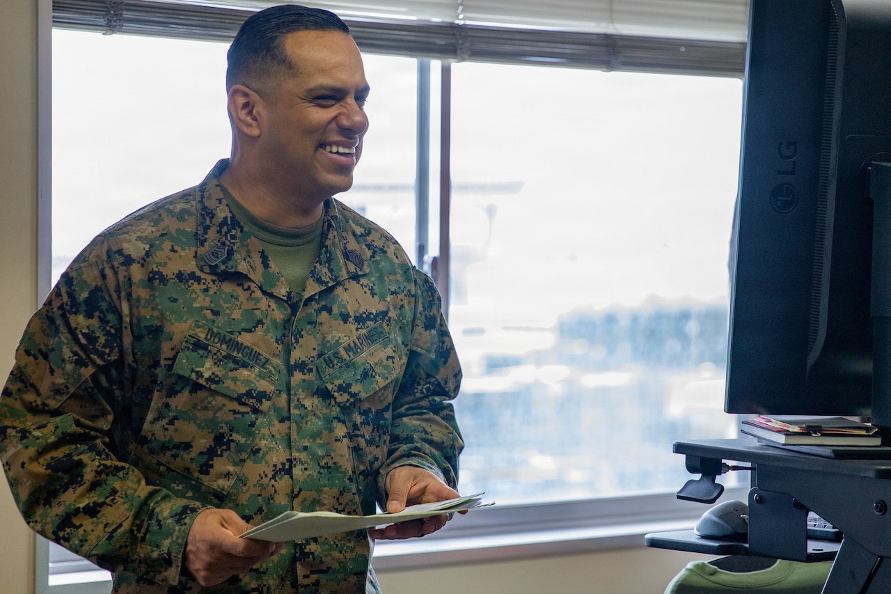 A Marine holding papers stands and smiles at something off-camera in an office-like setting, with a window in the background.