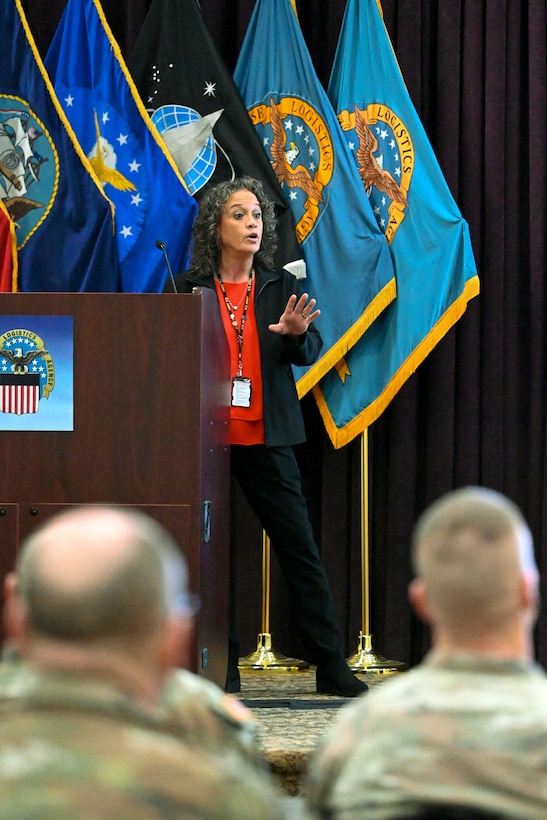 A woman in a dark suite stands on stage and talks to a room of people in military uniforms.