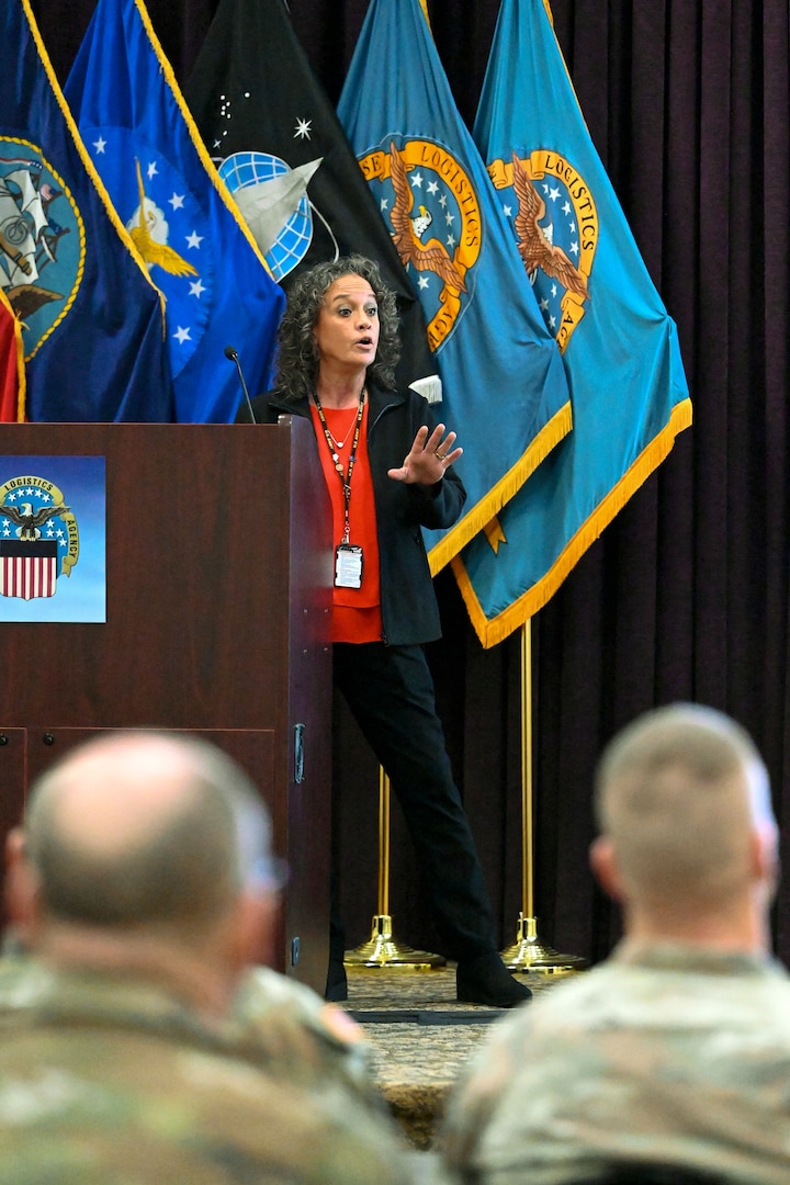 A woman in a dark suite stands on stage and talks to a room of people in military uniforms.